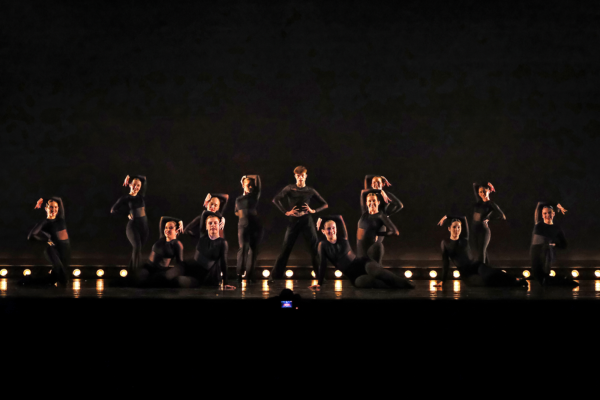 Group of dancers in black outfits posing on a dimly lit stage with warm floor lights. They strike dynamic poses in formation under stage lighting.
