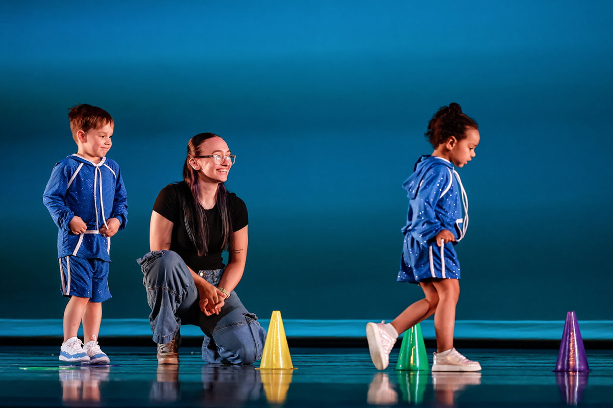 A smiling instructor kneels on the floor while two young children in blue athletic outfits walk past colorful cones on a stage-like floor.